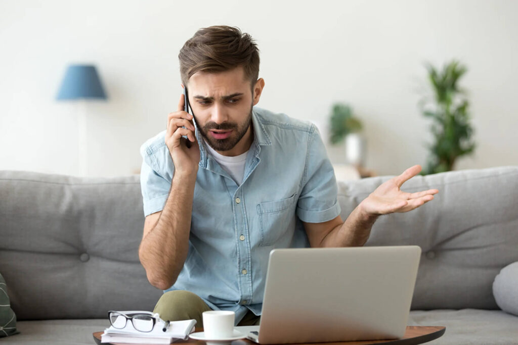 A man is sitting on a couch talking on a cell phone in front of a laptop computer.