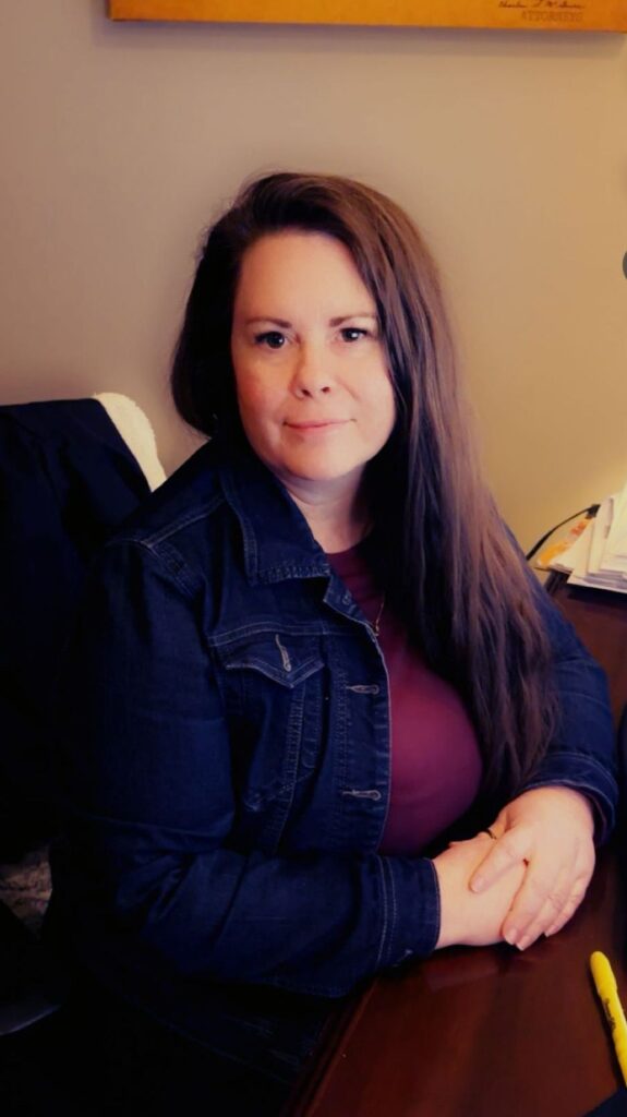 A woman in a denim jacket is sitting at a desk with her hands folded.