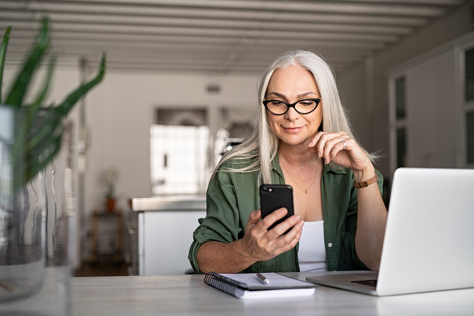 A woman is sitting at a desk using a laptop and a cell phone.
