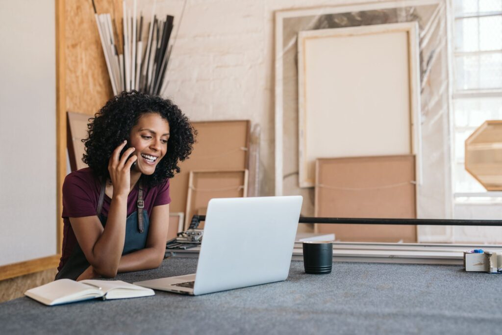A woman is sitting at a table with a laptop and talking on a cell phone.