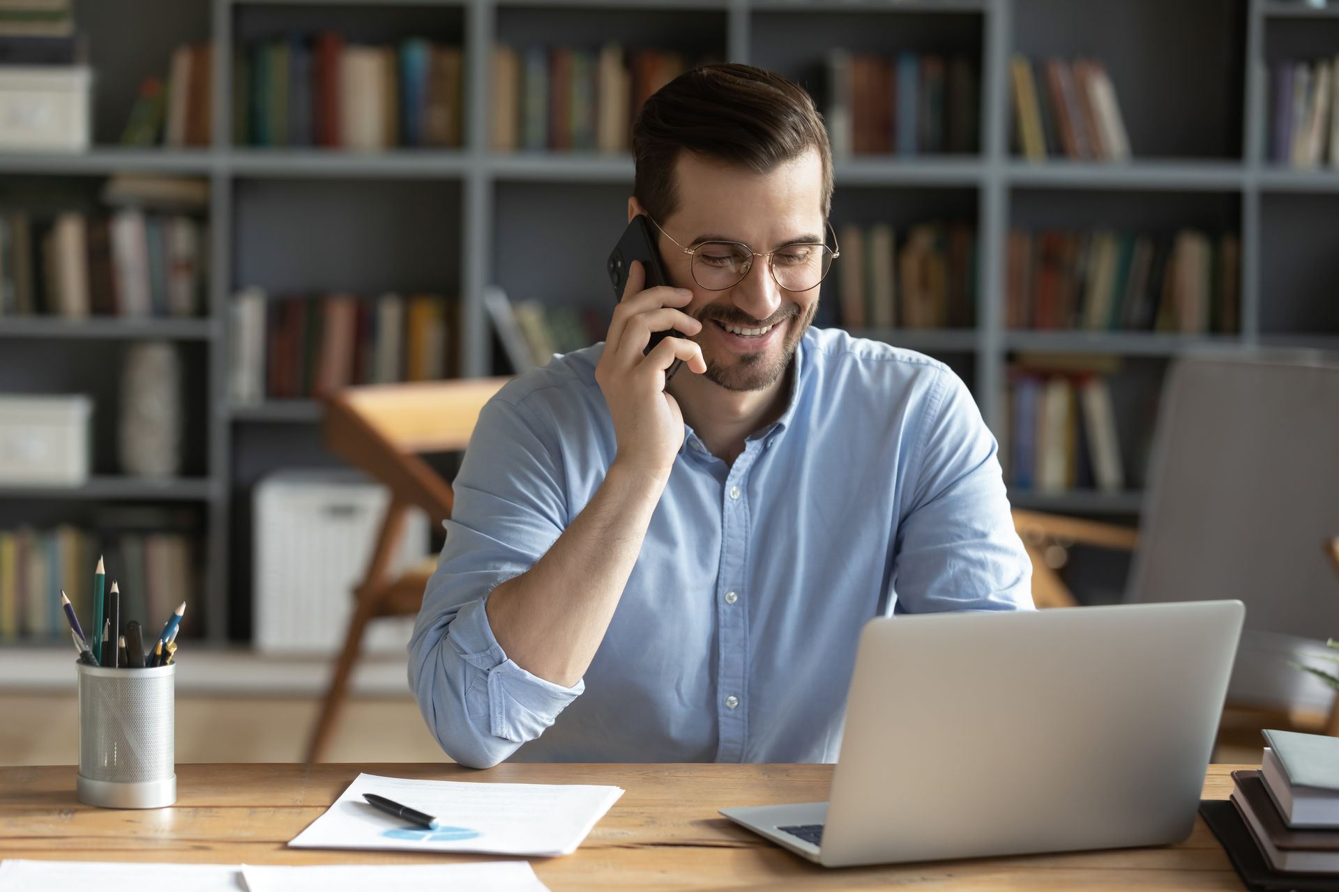 A man is sitting at a desk with a laptop and talking on a cell phone.