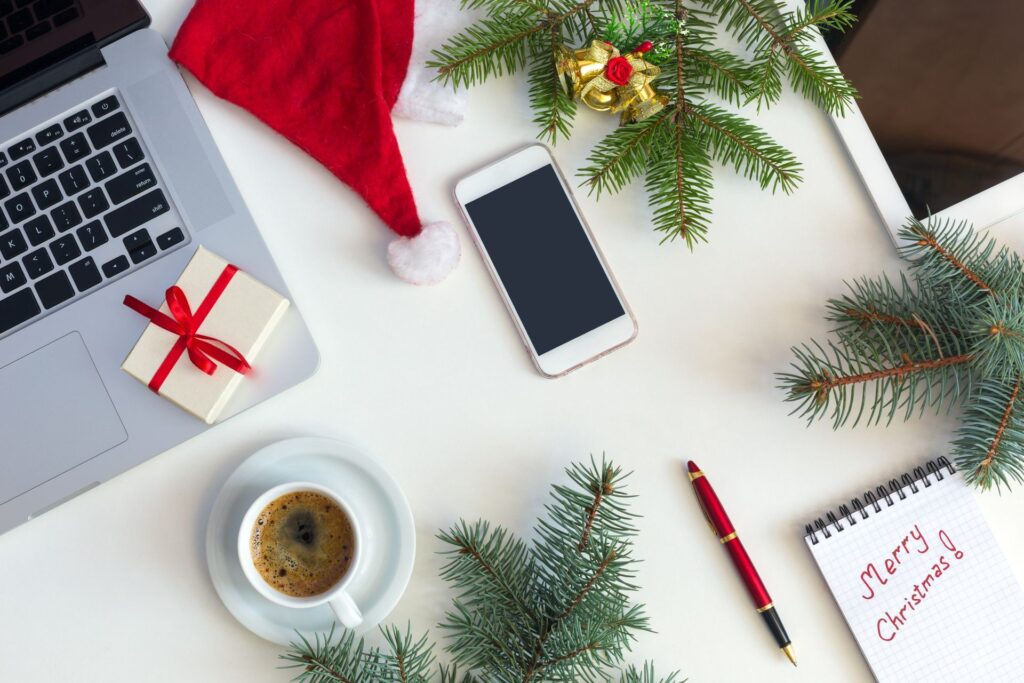 A desk with a laptop , a cell phone , a cup of coffee , a notebook and a santa hat.