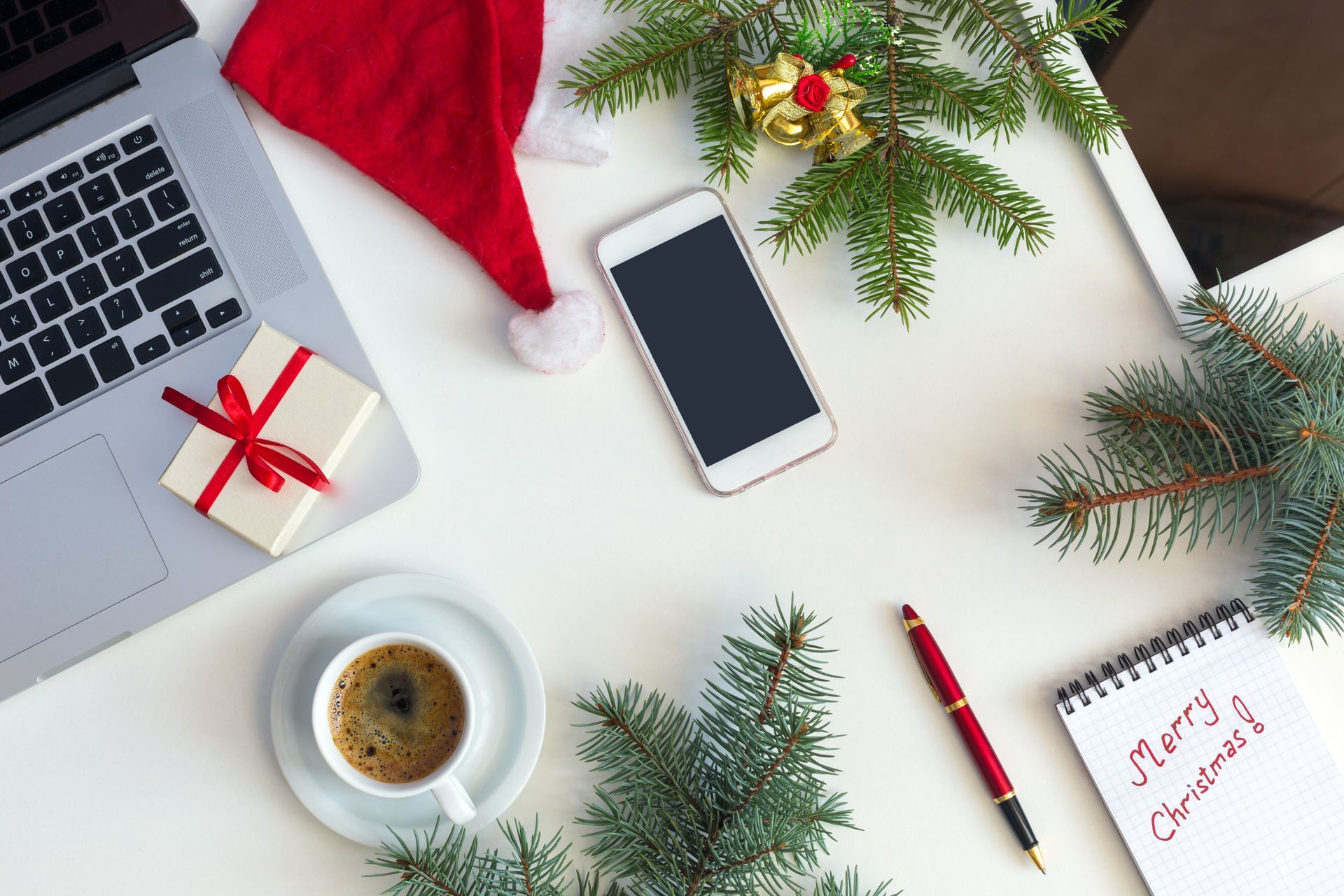 A desk with a laptop , a cell phone , a cup of coffee , a notebook and a santa hat.