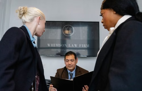 Three lawyers in a professional law firm boardroom with one man carefully reviewing a printed legal document at the table